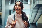 Junge Frau mit langen dunklen Haaren und rosa Pullover fasst an die Träger eines Rucksacks blickt nach links und lacht. Im Hintergrund Treppen und Fahrstühle.