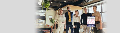 Fünf Kollegen stehen beieinander in einem Büro und lachen. Im Vordergrund das Kununu Top Company 2024 Logo.