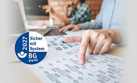 Ein Mann sitzt am Schreibtisch und zeigt auf ein Blatt Papier mit einem Diagramm, daneben das AMS-Güteseigel.