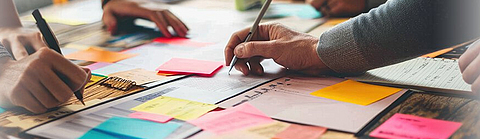 Verschiedene Menschen stehen um einen Tisch voller Dokumente und bunter Notizzettel und schreiben Ideen auf. | © MP Studio | Adobe Stock Verschiedene Menschen stehen um einen Tisch voller Dokumente und bunter Notizzettel und schreiben Ideen auf.